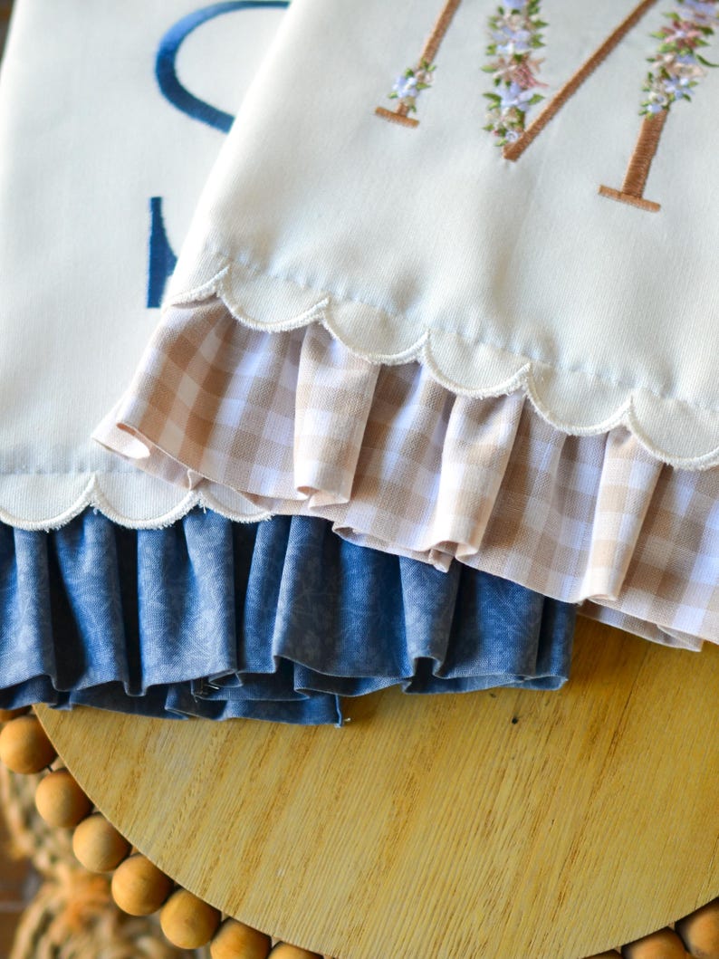 May include: Two off-white fabric towels with scalloped edges and ruffled trim. One towel features a brown embroidered letter M decorated with small floral accents, while the other displays a partial blue letter S. The ruffles are beige gingham.