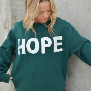 Puede incluir: Una sudadera verde oscuro con la palabra "HOPE" en letras grandes, blancas y texturizadas. La sudadera la lleva una persona con cabello rubio, contra una pared de hormigón gris. La prenda es informal y para uso diario.
