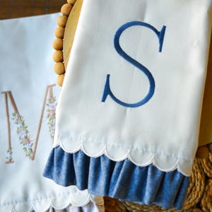 May include: Two white kitchen towels featuring scalloped edges and ruffled hems. One towel displays a large embroidered blue letter S, while the other features a large letter M decorated with small floral embroidery and a beige gingham ruffle.