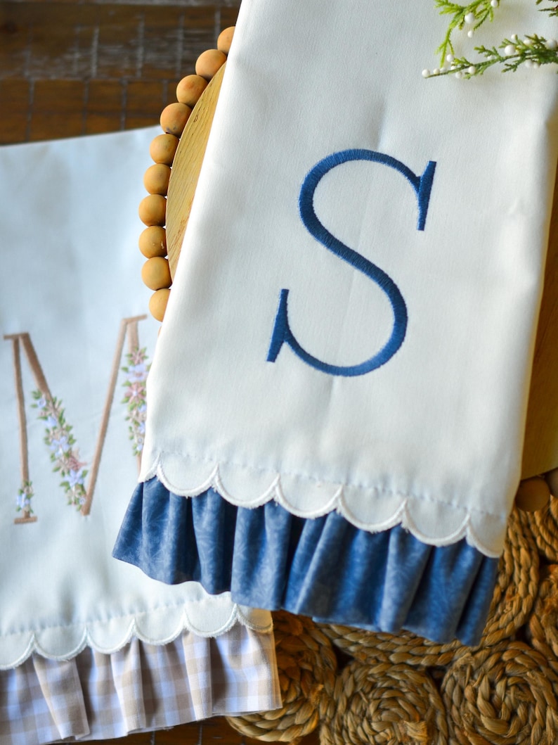 May include: Two white kitchen towels featuring scalloped edges and ruffled hems. One towel displays a large embroidered blue letter S, while the other features a large letter M decorated with small floral embroidery and a beige gingham ruffle.