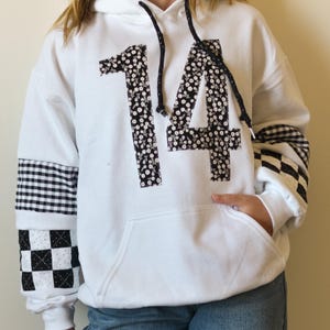 May include: White hoodie with the number "14" appliquéd in a black and white floral pattern. The sleeves feature black and white checkered and gingham patterns. The hoodie has a front pocket and black drawstrings.
