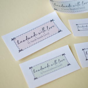 Op de afbeelding: Verschillende witte rechthoekige labels met de tekst "handmade with love" in een scriptlettertype. De labels hebben een geschulpte rand en strikaccenten. De labels zijn in verschillende maten en kleuren, waaronder roze en groen.