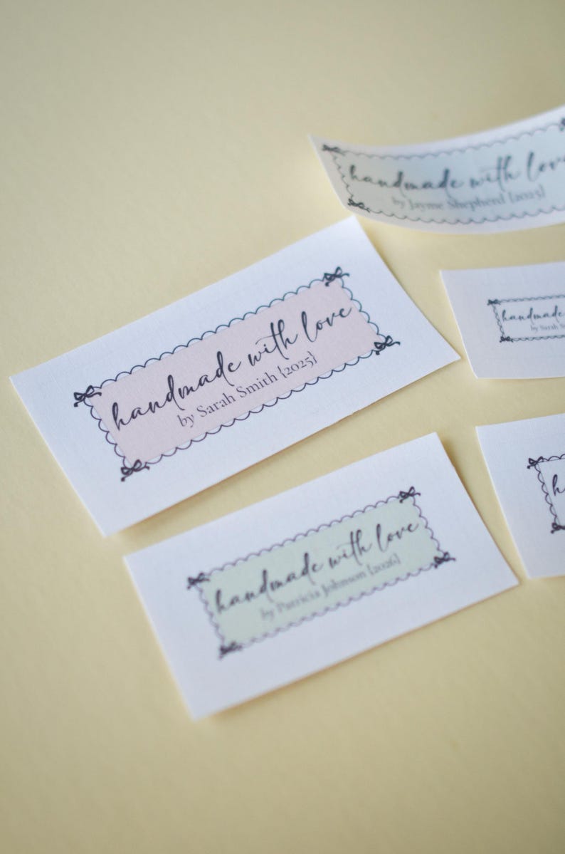 Op de afbeelding: Verschillende witte rechthoekige labels met de tekst "handmade with love" in een scriptlettertype. De labels hebben een geschulpte rand en strikaccenten. De labels zijn in verschillende maten en kleuren, waaronder roze en groen.