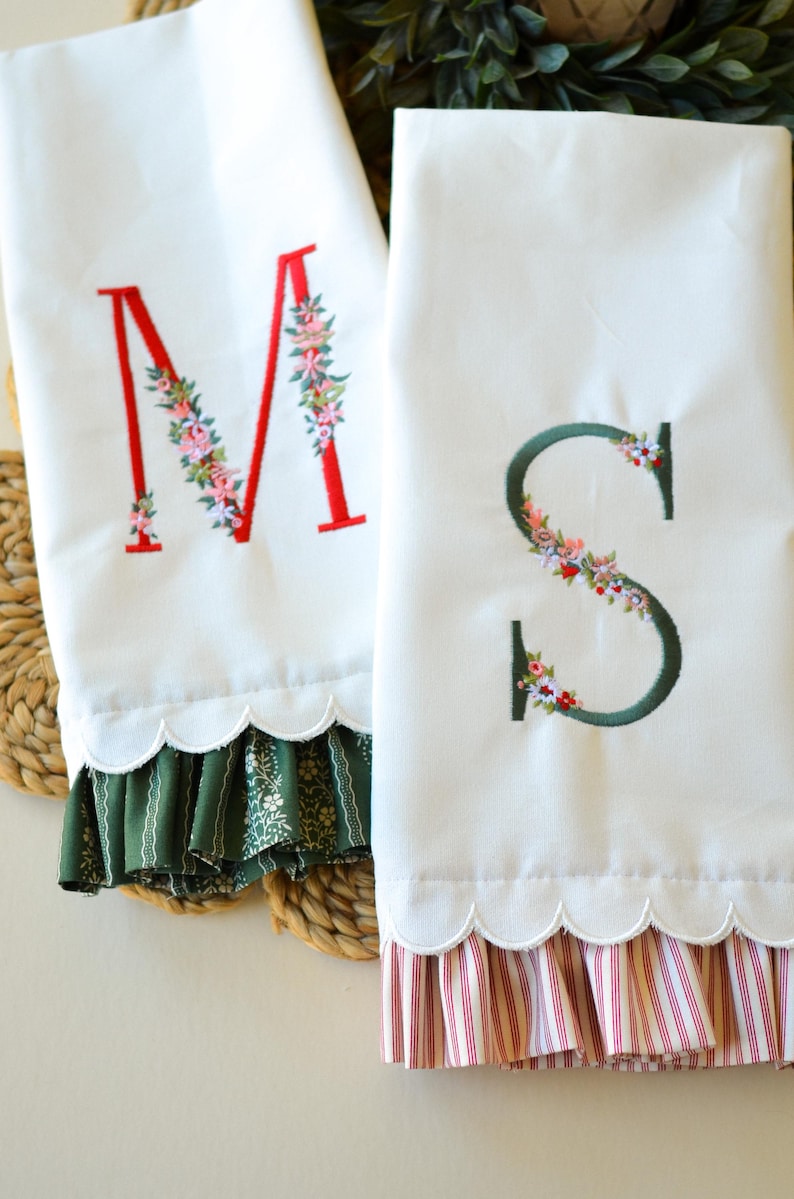 May include: Two white kitchen towels with scalloped edges and ruffled hems. One towel features a red embroidered letter M with floral accents, and the other features a dark green embroidered letter S with floral accents. The ruffles are patterned.