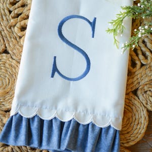 May include: A white hand towel featuring a large, embroidered blue letter S in the center. The bottom edge of the towel has a scalloped trim followed by a ruffled blue fabric border with a subtle tonal pattern. A small sprig of greenery is visible.