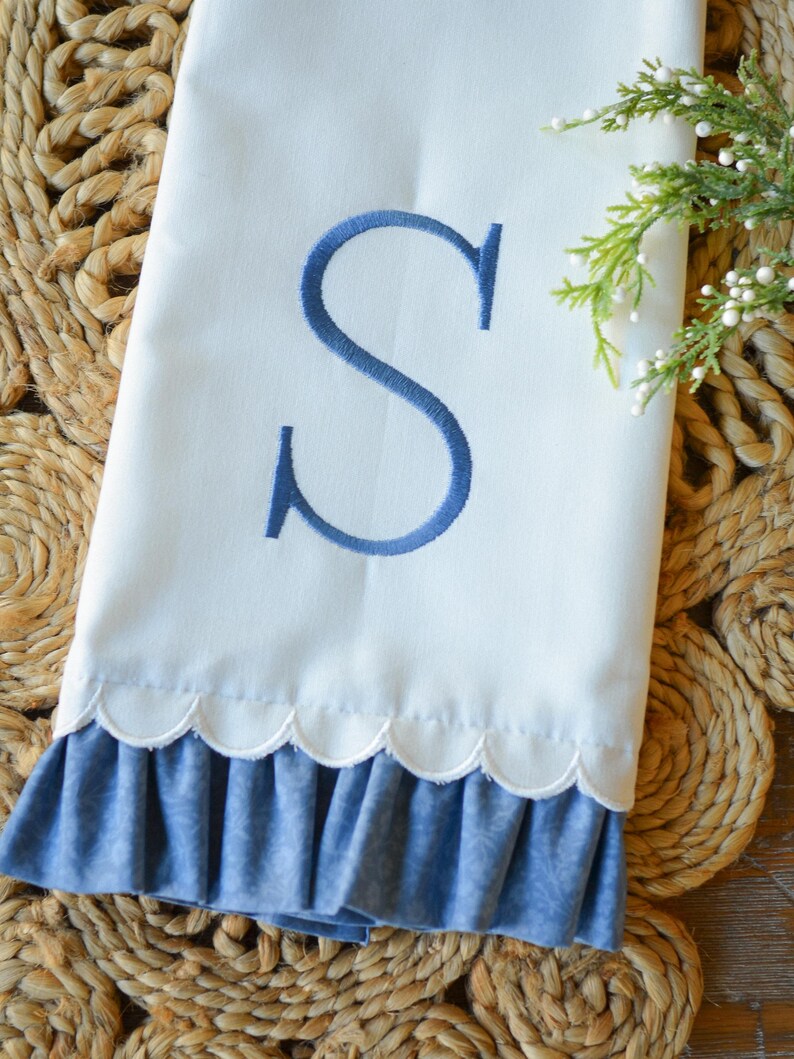 May include: A white hand towel featuring a large, embroidered blue letter S in the center. The bottom edge of the towel has a scalloped trim followed by a ruffled blue fabric border with a subtle tonal pattern. A small sprig of greenery is visible.