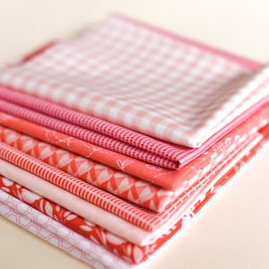 May include: A pile of folded fabric squares in shades of pink and red. The fabrics display various patterns, including gingham, polka dots, and geometric designs. The fabrics are neatly stacked, showcasing a variety of textures and colours.