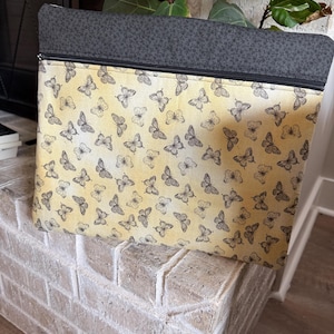 May include: Rectangular zipped pouch featuring a butterfly design on a yellow backdrop. The top section is dark grey fabric. The pouch rests on a white brick surface.