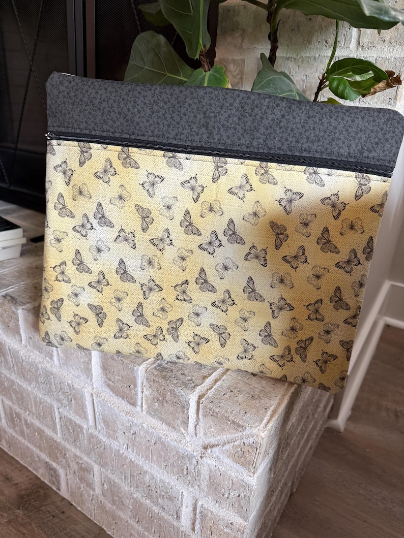 May include: Rectangular zipped pouch featuring a butterfly design on a yellow backdrop. The top section is dark grey fabric. The pouch rests on a white brick surface.
