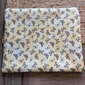 May include: A cream-coloured fabric featuring a repeating pattern of grey butterflies. The butterflies are spread across the surface, creating a delicate design. The fabric is likely used for crafting or home decor, and measures approximately 30 cm x 40 cm.