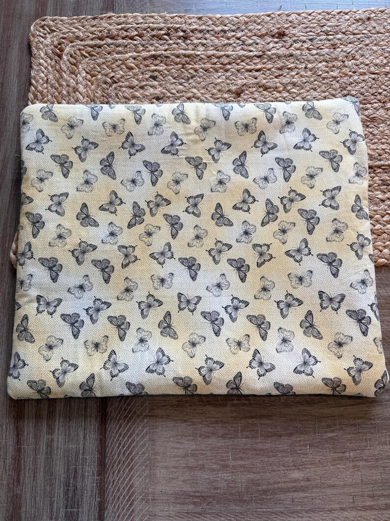 May include: A cream-coloured fabric featuring a repeating pattern of grey butterflies. The butterflies are spread across the surface, creating a delicate design. The fabric is likely used for crafting or home decor, and measures approximately 30 cm x 40 cm.