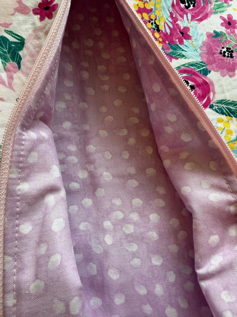 Op de afbeelding: Close-up van een bloemmotiefstof met een roze rits. De binnenvoering is lichtpaars met witte stippen. De buitenstof heeft roze en witte bloemen met groene bladeren.