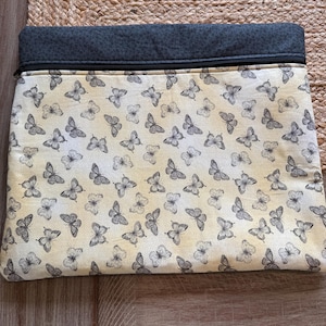 May include: A rectangular clutch bag featuring a cream-coloured background adorned with a pattern of grey butterflies. The top section is dark grey fabric, with a black zip. The bag rests on a woven, tan surface.
