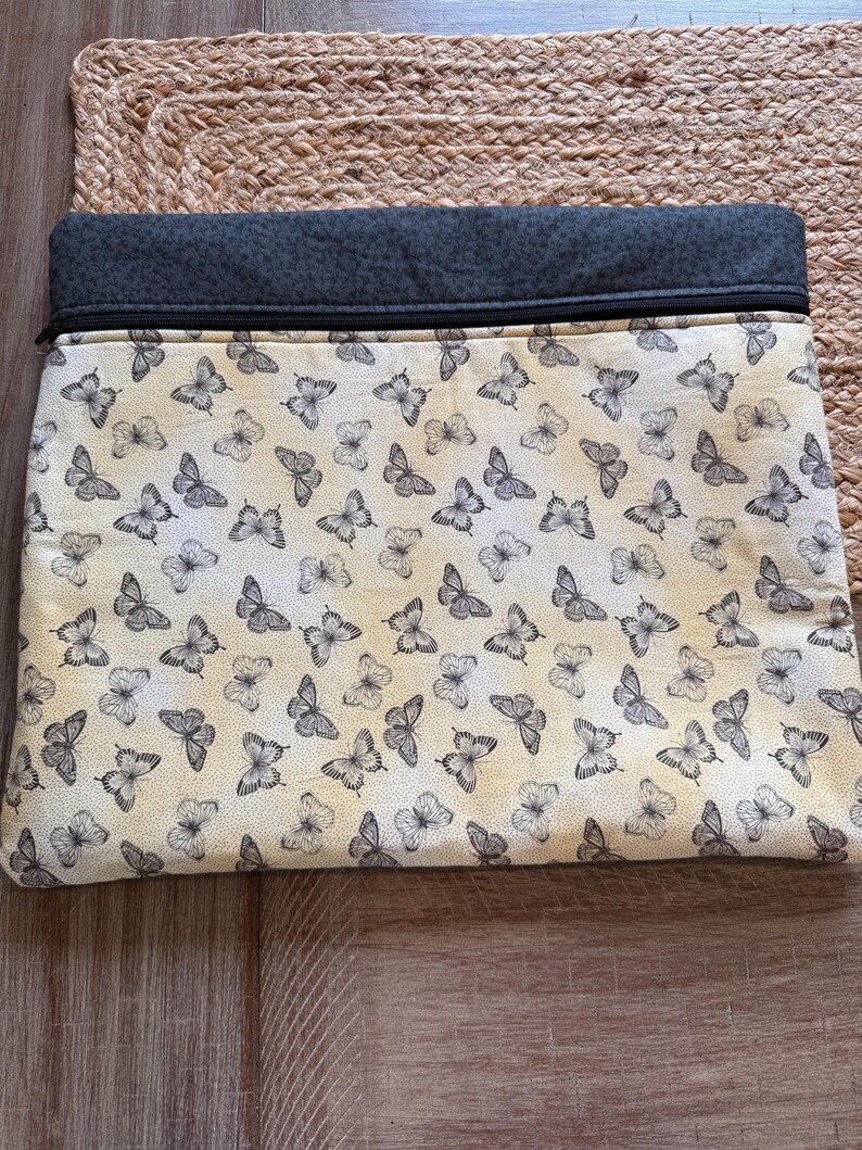 May include: A rectangular clutch bag featuring a cream-coloured background adorned with a pattern of grey butterflies. The top section is dark grey fabric, with a black zip. The bag rests on a woven, tan surface.