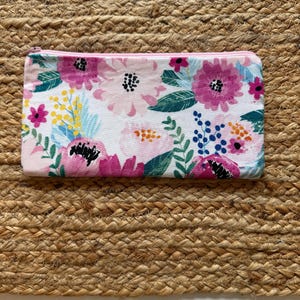 Op de afbeelding: Een rechthoekig etui met een bloemenpatroon en een roze rits. Het etui heeft een witte achtergrond met roze, paarse en gele bloemen, en groene bladeren. Het ligt op een geweven, bruin oppervlak.