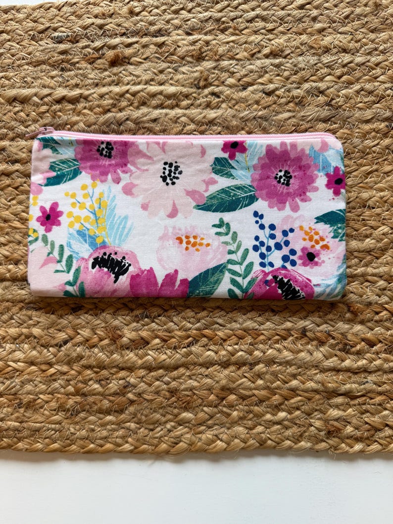 Op de afbeelding: Een rechthoekig etui met een bloemenpatroon en een roze rits. Het etui heeft een witte achtergrond met roze, paarse en gele bloemen, en groene bladeren. Het ligt op een geweven, bruin oppervlak.
