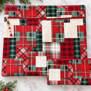 May include: A large rectangular fabric pouch and a smaller matching rectangular fabric pouch, both featuring a patchwork design of red, green, and cream plaid patterns. Each pouch has a red zipper closure with a beaded pull on the larger one.