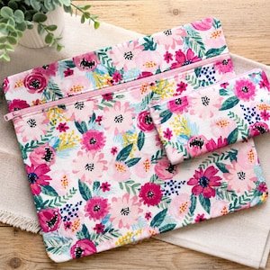 Op de afbeelding: Twee etuis met ritssluiting en een bloemenpatroon. De etuis hebben een witte achtergrond met roze, fuchsia en gele bloemen en groene bladeren. De ritsen zijn roze. De etuis liggen op een witte doek.