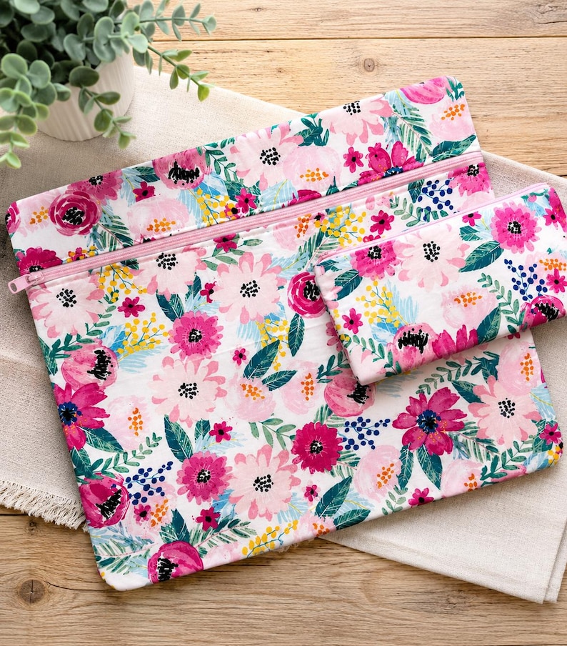 Op de afbeelding: Twee etuis met ritssluiting en een bloemenpatroon. De etuis hebben een witte achtergrond met roze, fuchsia en gele bloemen en groene bladeren. De ritsen zijn roze. De etuis liggen op een witte doek.