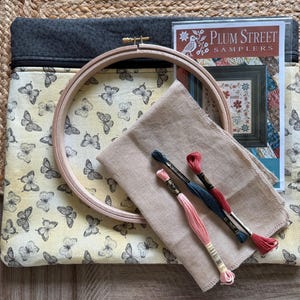 May include: A craft kit with a butterfly-patterned case, a wooden embroidery hoop, fabric, and embroidery floss in pink, teal, and red. A booklet titled "Plum Street Samplers" is included. The case has a grey top and a zip closure.