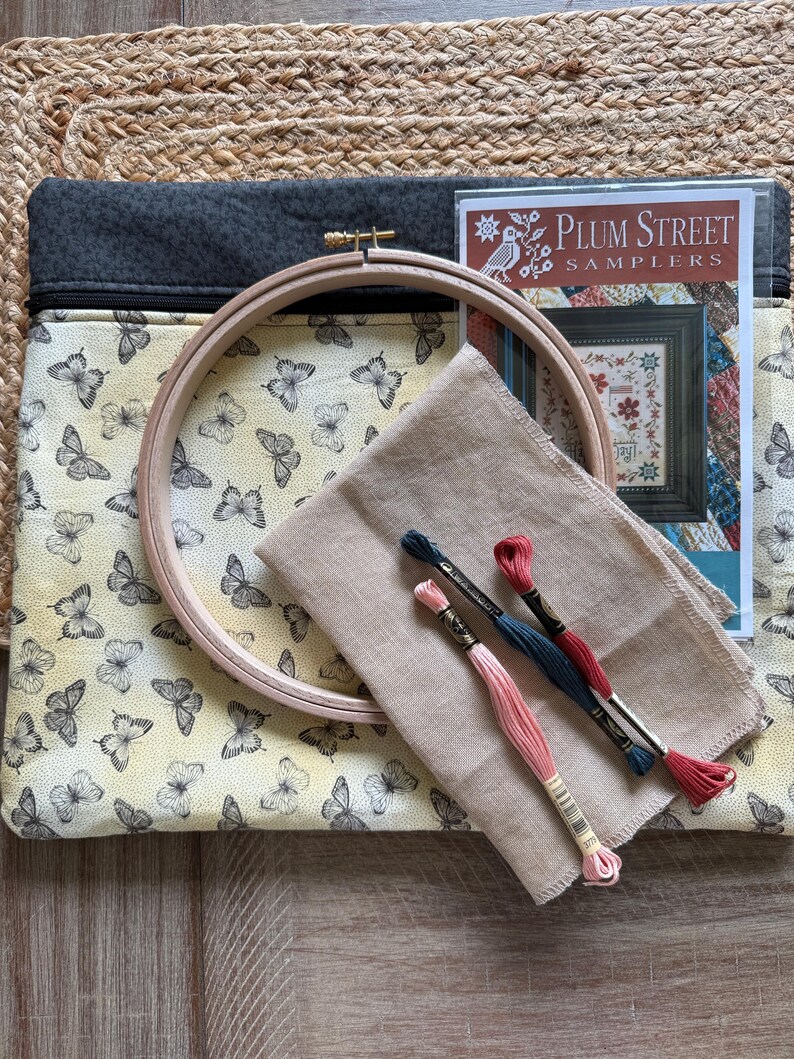 May include: A craft kit with a butterfly-patterned case, a wooden embroidery hoop, fabric, and embroidery floss in pink, teal, and red. A booklet titled "Plum Street Samplers" is included. The case has a grey top and a zip closure.