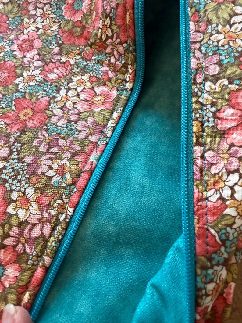 Pode incluir: Close-up de um tecido com padr&atilde;o floral com um fecho de correr turquesa. O tecido apresenta um fundo castanho com flores rosa, vermelhas, brancas e azuis. O forro interior &eacute; de cor turquesa lisa.