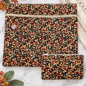May include: A large rectangular fabric pouch and a smaller matching rectangular fabric pouch, both featuring a dense pattern of autumn leaves in shades of orange, yellow, red, and green on a black background, with gold zippers and a leaf charm.