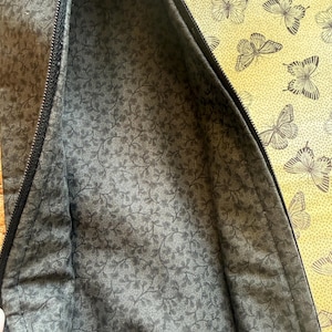 May include: A partially open zippered pouch with a dark grey floral patterned interior. The exterior features a yellow fabric with a repeating butterfly design. The zipper is black.