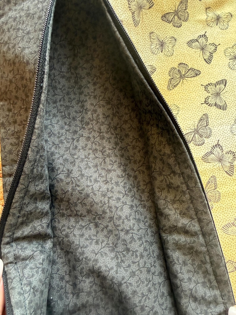 May include: A partially open zippered pouch with a dark grey floral patterned interior. The exterior features a yellow fabric with a repeating butterfly design. The zipper is black.