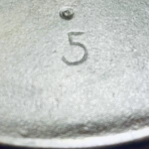May include: Close-up of a well-worn, black cast iron skillet. The number "5" is prominently displayed on the cooking surface. The handle is visible, and the skillet rests on a red and white checkered cloth.