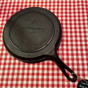 May include: A black cast iron griddle with a handle and a circular cooking surface. The griddle is 8 1/2 inches in diameter. The griddle is on a red and white checkered tablecloth. A tag reads "Late 19th Gate Mark Griddle 8 1/2".