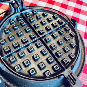 May include: A round, black cast iron waffle maker featuring a classic grid pattern with deep, square indentations. The cooking surface is divided into four distinct quadrants by a cross-shaped divider, showing a textured, metallic finish.