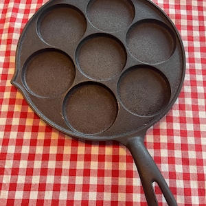 May include: A cast iron pan with seven circular indentations for cooking. The pan has a long handle with a hole for hanging. The pan is dark brown and sits on a red and white checkered tablecloth.