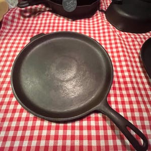 May include: A black cast iron griddle pan with a long handle and a round cooking surface. The pan is sitting on a red and white checkered tablecloth. Other cast iron cookware is visible in the background.