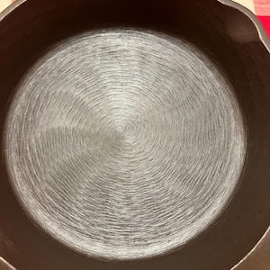 May include: A dark gray cast iron skillet featuring a circular, machined texture on the interior cooking surface. The pan has a smooth, rounded rim and a distinct pouring spout on the upper right side, showcasing a metallic, brushed finish.
