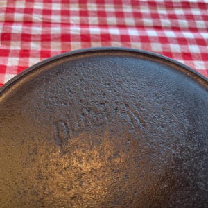 May include: Close-up of a dark, textured cast iron skillet. The word "DUSE" is embossed on the surface. The skillet is set against a red and white checkered tablecloth.
