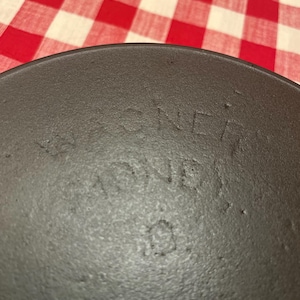 May include: Close-up of a dark gray cast iron pan with the word "WAGNER" embossed on the surface. The pan has a textured surface and a slightly curved edge. The background is a red and white checkered pattern.