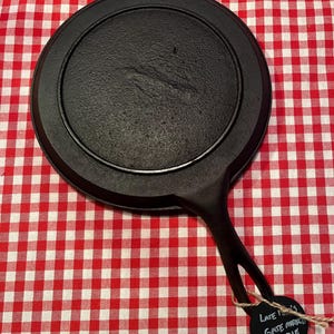 May include: A black cast iron griddle with a single handle. The griddle is round with a raised edge and a textured cooking surface. A tag attached to the handle reads "Late 1900's Gate Marked Griddle 8 1/2".