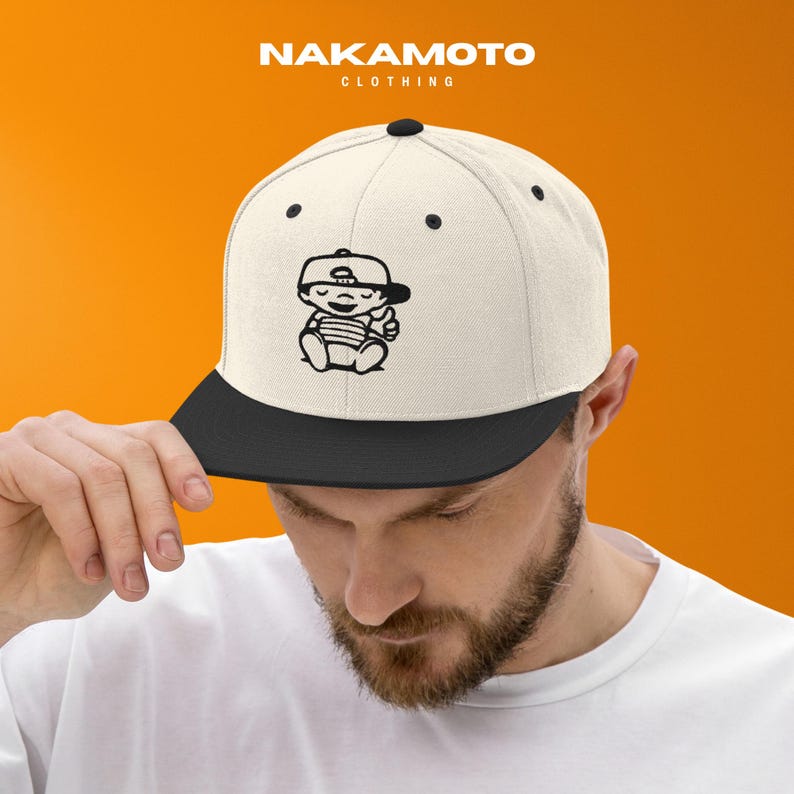 Puede incluir: Gorra plana blanca y negra con un dibujo de un beb&eacute; de dibujos animados. La gorra tiene una visera negra y una corona blanca. El dibujo del beb&eacute; es negro y muestra a un beb&eacute; con gorra haciendo una se&ntilde;al de pulgar hacia arriba. La marca "NAKAMOTO CLOTHING" est&aacute; impresa encima.