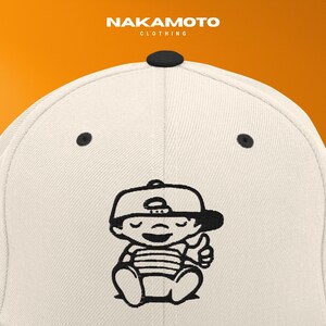 Puede incluir: Gorra de b&eacute;isbol crema con visera negra y un dibujo animado negro de un beb&eacute; haciendo un gesto de pulgar hacia arriba. El nombre de la marca "NAKAMOTO CLOTHING" est&aacute; impreso en la parte superior. Un accesorio de moda.