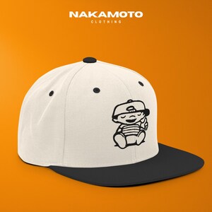 Puede incluir: Gorra de b&eacute;isbol blanca y negra con visera plana. La gorra presenta un gr&aacute;fico de dibujos animados en blanco y negro de un beb&eacute; con gorra. El texto "NAKAMOTO CLOTHING" est&aacute; en la parte superior.