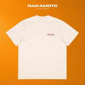 Puede incluir: Camiseta color crema con la palabra "HODL" en rojo, centrada en el pecho. La camiseta tiene cuello redondo y mangas cortas. La marca "NAKAMOTO CLOTHING" está impresa en blanco en la parte superior.