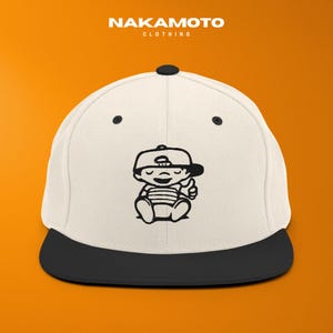 Puede incluir: Gorra de b&eacute;isbol blanca y negra con visera negra. La gorra presenta una ilustraci&oacute;n de dibujos animados en blanco y negro de un beb&eacute; haciendo una se&ntilde;al de pulgar hacia arriba. El texto "NAKAMOTO" est&aacute; en la parte superior.