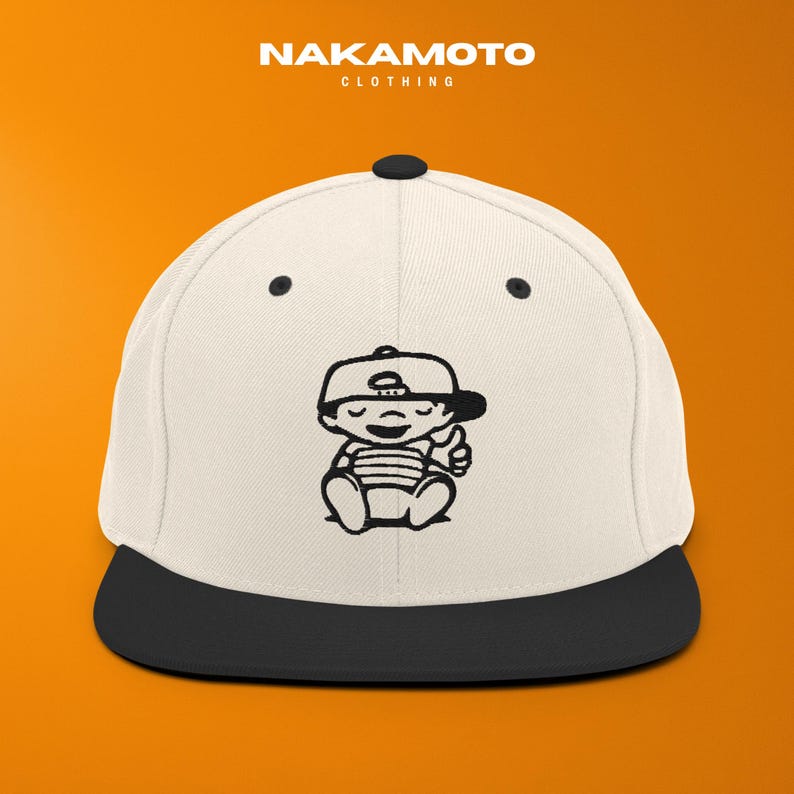 Puede incluir: Gorra de b&eacute;isbol blanca y negra con visera negra. La gorra presenta una ilustraci&oacute;n de dibujos animados en blanco y negro de un beb&eacute; haciendo una se&ntilde;al de pulgar hacia arriba. El texto "NAKAMOTO" est&aacute; en la parte superior.