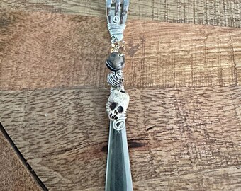 May include: A decorative fork with a skull and bead embellishments. The fork's tines are at the top, with wire wrapping and decorative beads in the middle. A silver-colored skull is at the base of the fork.