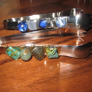 May include: Two pairs of silver serving tongs with decorative wire wrapped beads. The top pair has blue and white beads. The bottom pair has turquoise, green, and brown beads.