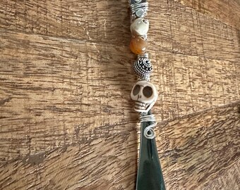 May include: A silver-toned shoe horn with a decorative handle. The handle features a series of beads, including skulls, an orange bead, and a silver-colored bead. The shoe horn is on a wooden surface.