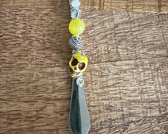 May include: A silver shoe horn with a decorative handle. The handle features yellow and silver beads, and a yellow skull. The shoe horn is on a wooden surface.