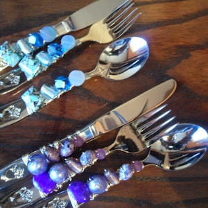 May include: Set of four silver utensils decorated with colorful beads. The utensils include a knife, fork, and two spoons. The beads are blue, purple, and silver.