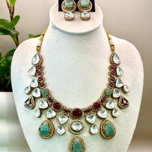 May include: A gold-toned necklace and earring set. The necklace features clear, teardrop-shaped stones, red stones, and green stones. The earrings have a similar design. The jewelry is displayed on a white bust.